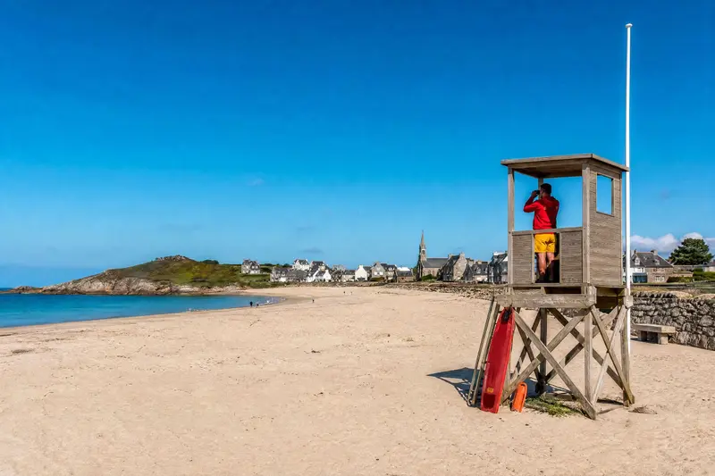 Poste de secours en bois sur pilotis en haut de la plage, un sauveteur en rouge observe la mer aux jumelles ; le mât est vide.