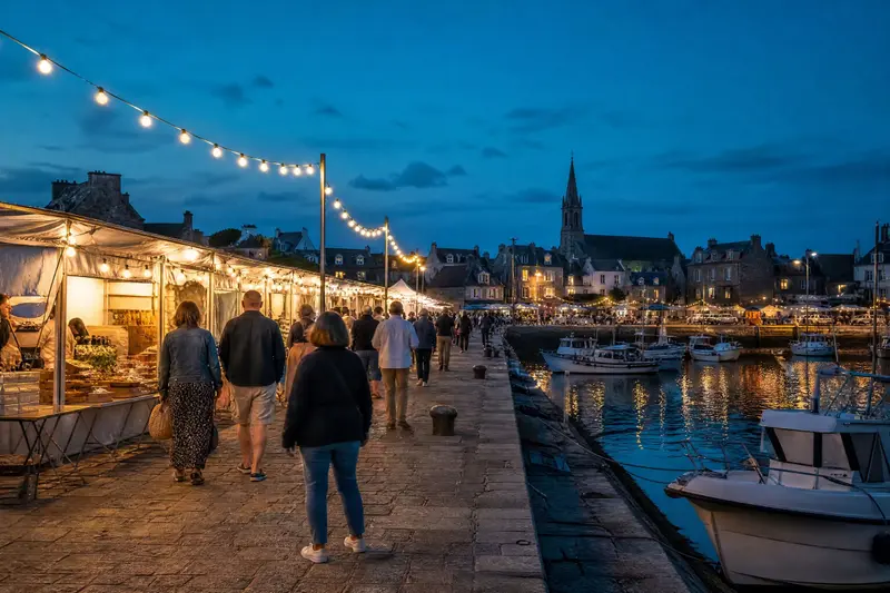 Le marché nocturne sur le quai : stands blancs sous des guirlandes lumineuses, promeneurs de dos.