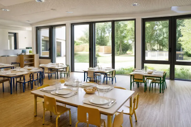 Le réfectoire de l'école, vide, aux grandes fenêtres : tables basses dressées et petites chaises de couleur.